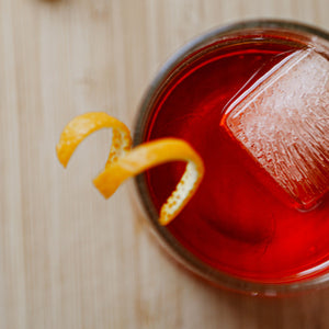 Downpour Oak Aged Negroni. A glass of bright orange red liquid, with a square ice cube and a twist of orange peel 