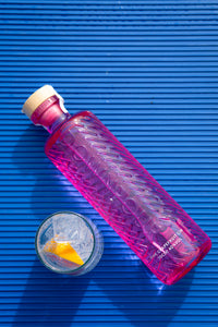Downpour Pink Grapefruit Gin, a bright pink bottle with a wooden stopper, lying on a blue lined background. 