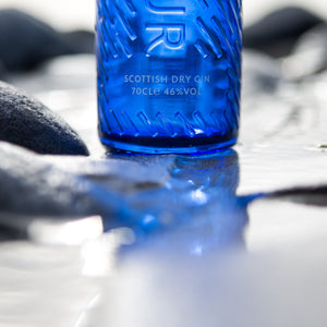 A close up of Downpour Scottish Dry Gin, in a blue glass bottle, sitting on wet sand