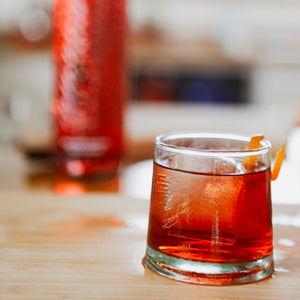 A glass of Negroni, with ice and a twist of orange peel. It is sitting on a wooden chopping board and out of focus in the back ground is a bottle of the same orangey red liquid. 