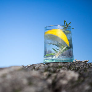 Downpour Perfect Serve. A short glass of G&T, with a wedge of lemon, ice, and a sprig of rosemary. The glass is sitting on a rock against a bright blue sky.